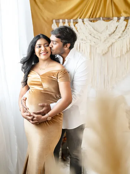 A sweet and affectionate moment captured in a bright, boho-style room. The partner embraces the mom-to-be from behind, kissing her cheek as she smiles, both celebrating the baby on the way.