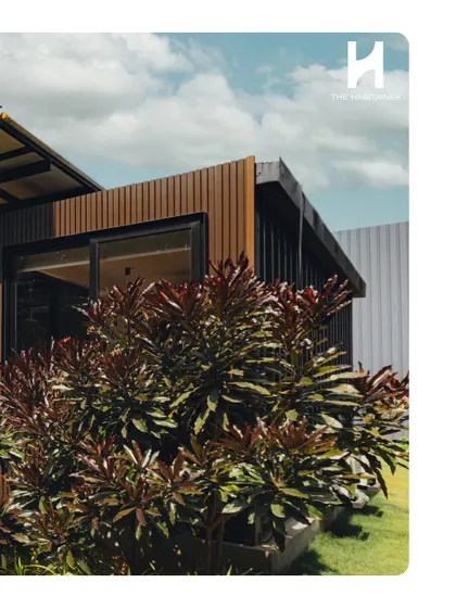 A detail shot of the corner of the twin container office, showing the intersection of the wood-look vertical cladding and the large window, framed by lush plants.
