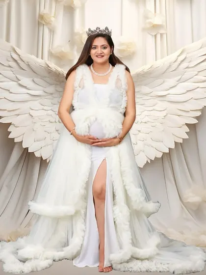 A powerful and angelic studio portrait of an expectant mother wearing a beautiful white gown and a crown. She stands gracefully with large, white angel wings behind her against a dreamy, cloud-like backdrop, portraying the miracle of motherhood.