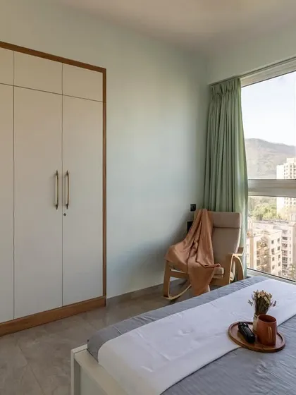 This bedroom with a large window and stunning city view is kept minimal and airy with a simple white and wood wardrobe and a comfortable rocking chair.