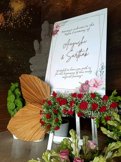 A personalized welcome sign for Aayushi and Sarthak's Roka ceremony. The elegant design is complemented by red roses and a serene Buddha statue in the background.