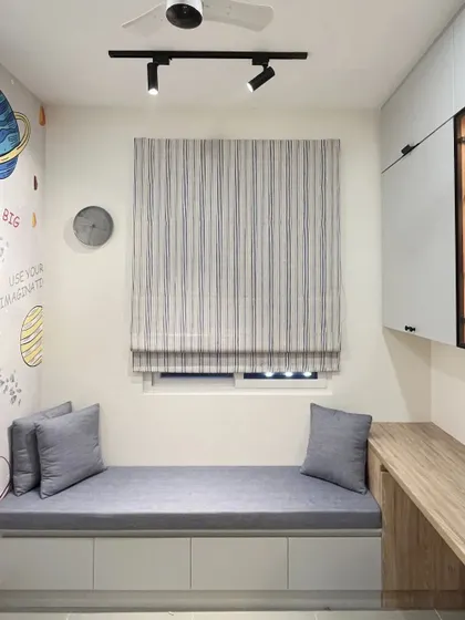 A simple and clean bay window seat with grey upholstery and under-seat storage, located next to a study desk in a kid's room.