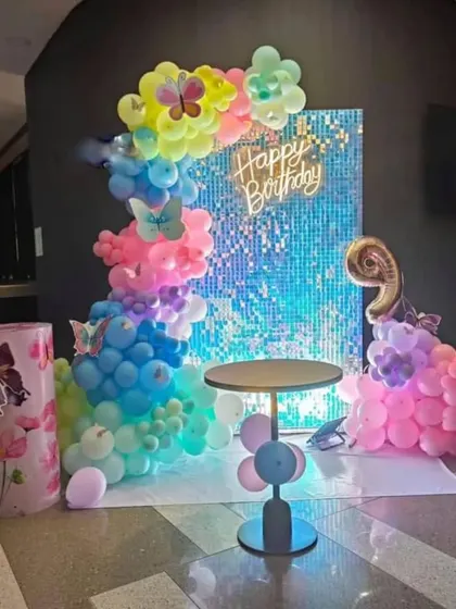 A modern butterfly-themed party with a shimmering blue sequin backdrop. The decoration features a rainbow balloon garland, a neon sign, and a custom-decorated pedestal.