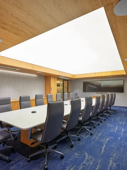 A large, formal boardroom featuring a stretch-lit ceiling that provides bright, even, and glare-free light. The room is equipped with a long conference table, comfortable seating, and an additional bench along the wall for larger meetings.
