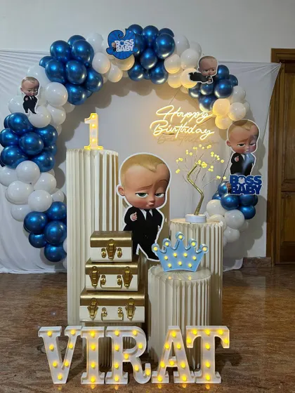 A fun Boss Baby themed first birthday party. The decoration features a blue and white balloon arch, character cutouts, marquee letters for the name, and a 'Happy Birthday' neon sign.