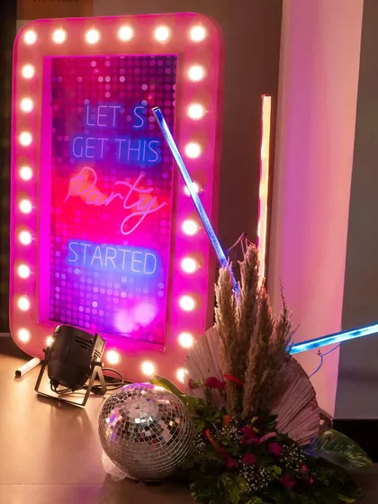 A vibrant neon sign that reads 'Let's Get This Party Started', framed with pink marquee lights and a disco ball, setting the mood for a disco party.