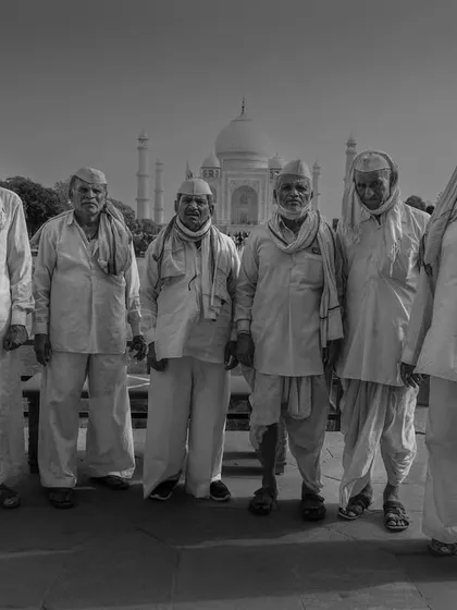 A group of elderly men in traditional attire stand for a portrait in front of the Taj Mahal. This black and white photo captures a moment of cultural juxtaposition.