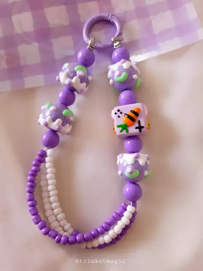 This purple and white beaded charm features fun, cake-like beads and a cute character cube, adding a playful and unique touch to your phone or bag.