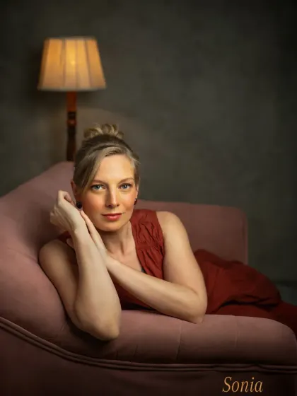 A serene and confident portrait of Sonia, resting on a pink chaise lounge. The soft lighting from the antique lamp and the moody backdrop create an intimate, reflective atmosphere.