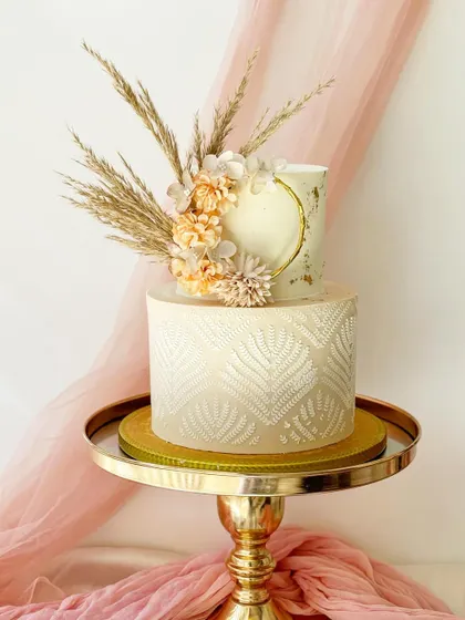 An elegant two-tier cake perfect for a modern celebration. The bottom tier features a chic white leaf stencil pattern, while the top is accented with gold leaf and topped with dried pampas grass.