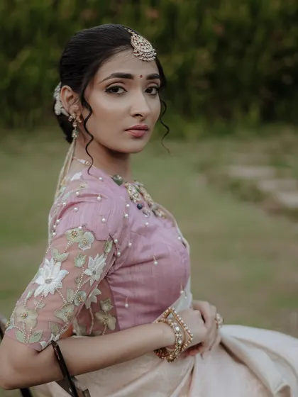 A classic and elegant portrait of a bride seated outdoors, looking gracefully towards the camera. Her embroidered blouse and serene expression are the focus.