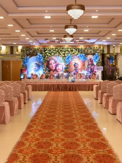 The event hall is set up for a first birthday party, with a long aisle leading to the Krishna-themed stage.