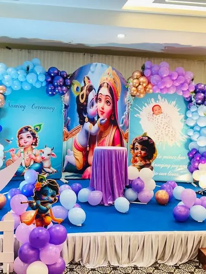 A beautiful Krishna-themed baby naming ceremony. The stage features character backdrops, a crescent moon prop, and balloon arrangements in shades of purple and blue.