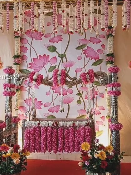 A beautiful naming ceremony design featuring a Pichwai-style lotus painting as the backdrop. The cradle is decorated with pink flowers, and the entire setup is framed by hanging jasmine garlands.