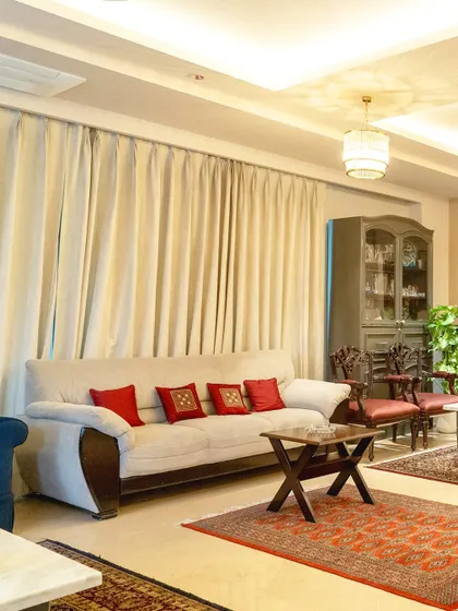 This cozy corner of the living room balances classic and contemporary elements. A neutral sofa is accented with vibrant red cushions, while traditional carved chairs and oriental rugs ground the space in timeless style.