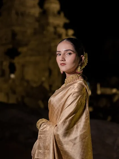 A portrait of a bride, bathed in the soft glow of night. The dramatic lighting against the temple backdrop creates a powerful and soulful image, showcasing her poise and the richness of her golden saree.