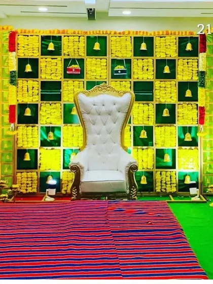 A bright and traditional Seemantham stage with a green and yellow checkerboard backdrop. The decor includes hanging bells and is set up with a traditional woven mat for the ceremony.