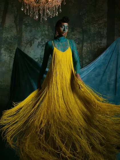 A fine art fashion portrait combining a dramatic, fringed dress with conceptual makeup and a painterly background.