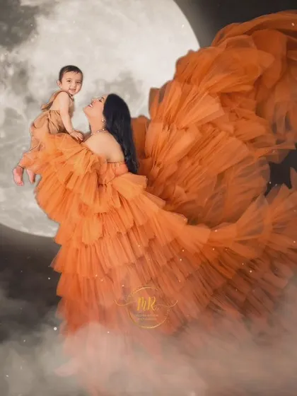 A creative "over the moon" concept for a mother and baby portrait. The flowing orange gown and moon backdrop create a magical and whimsical scene.