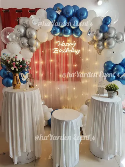 A blue, white, and silver balloon arch framing a set of tables with a fairy light curtain in the background.