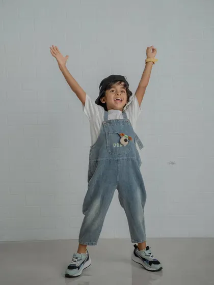 An energetic shot of a boy in overalls and a cap with his arms raised in excitement against a white brick wall.