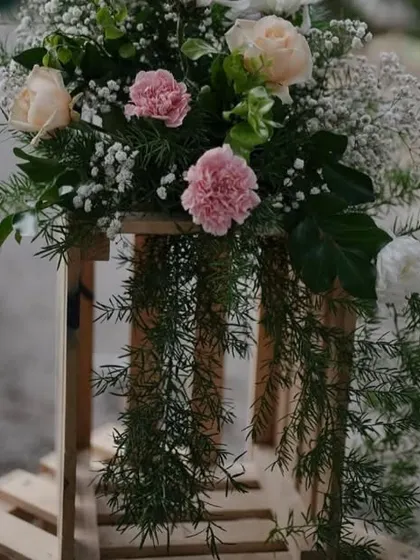 A rustic floral arrangement on a wooden crate. The combination of soft pink carnations, white roses, and cascading greenery creates a natural and romantic feel.