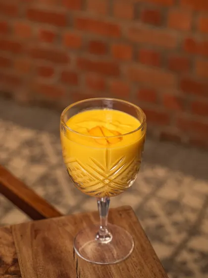 A thick mango smoothie in a crystal glass, shot against a rustic brick wall. The simple but elegant presentation highlights the drink's rich texture and color.