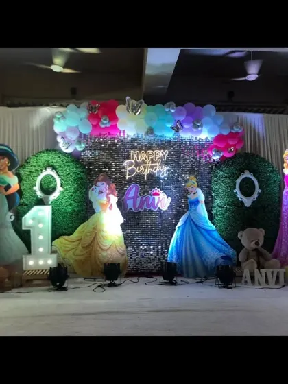 A Disney princess themed first birthday stage, with cutouts of various princesses, a green hedge backdrop, and a light-up number '1'.