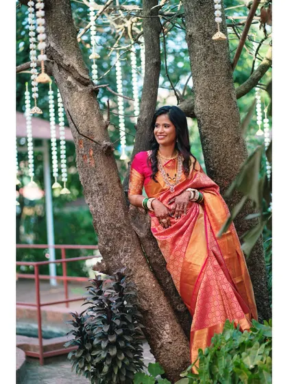 The bride looking radiant in her pink and gold saree, posing gracefully under a tree decorated with traditional hangings for her engagement.