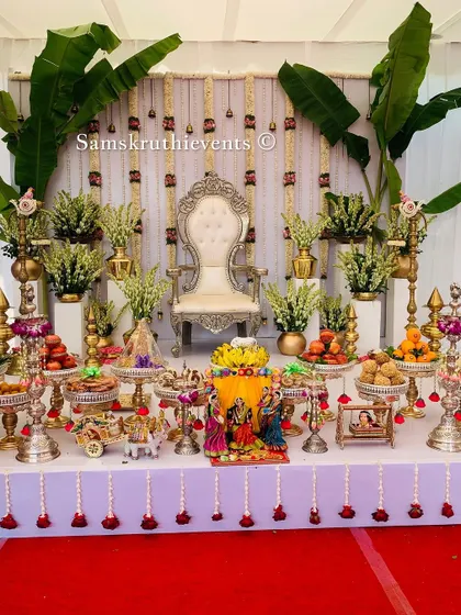 This white and gold themed setup brings a touch of serene elegance to the Seemantha ceremony. The stage is adorned with banana leaves and tuberose flower hangings, complemented by a full 21-plate arrangement on silver stands.