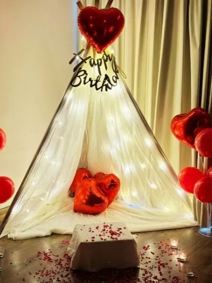 A romantic and intimate birthday surprise created right in the bedroom. This cozy setup features a beautiful teepee tent draped in sheer fabric and fairy lights, with red heart-shaped balloons and scattered rose petals to create a special, personal celebration.