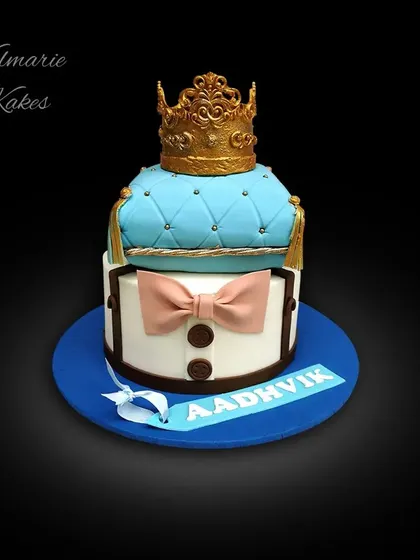 A royal-themed cake fit for a little prince. This design features a quilted blue pillow topped with a golden crown, all made from fondant, with a cute bow tie detail on the bottom tier.