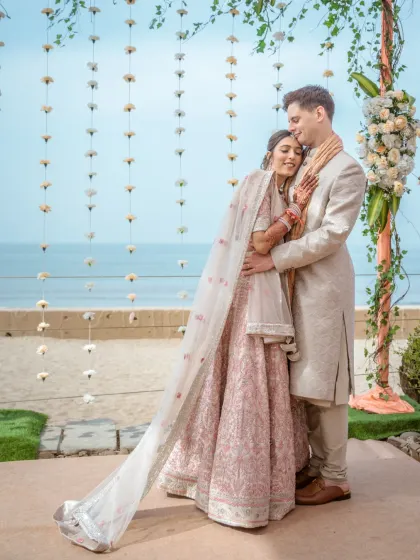 A full-length portrait of the couple embracing by the sea. The bride's flowing dupatta and the serene ocean background create a graceful and elegant composition.