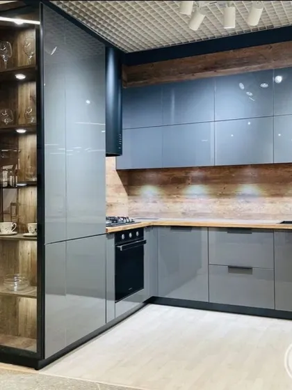 This design pairs glossy grey handleless cabinets with a warm wood-textured backsplash and countertop. A tall glass-fronted unit provides elegant display storage, creating a balanced and modern kitchen.