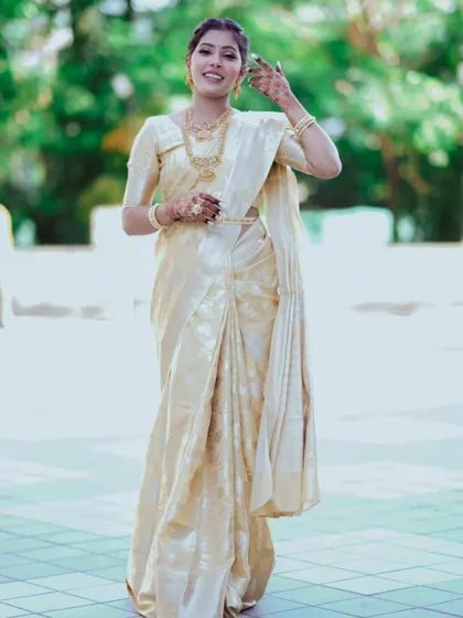 A full-length portrait of a woman in an elegant off-white saree, smiling warmly. The focus is on her complete traditional look, from her jewelry to her confident posture.