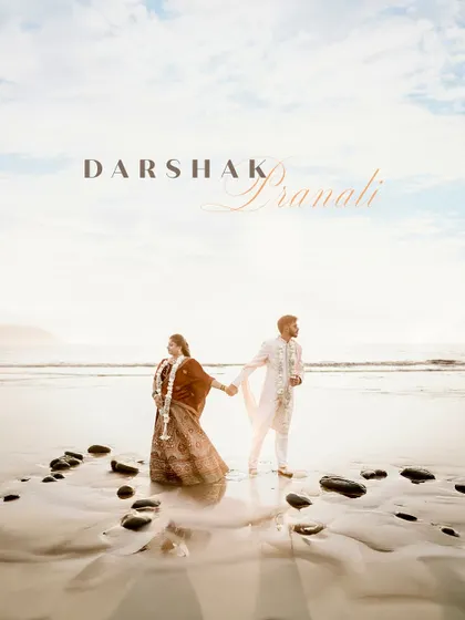 A breathtaking destination wedding portrait on the beach. The couple holds hands against a vast ocean and a beautiful sky, creating a truly epic and romantic memory of their special day.