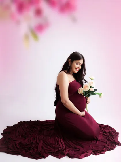 A soft and romantic portrait. The mom-to-be kneels in a flowing burgundy gown, holding a small bouquet of roses against a gentle pink backdrop.