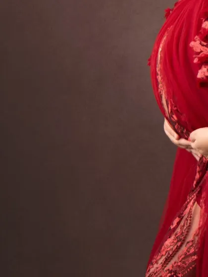 A beautiful close-up detail shot, focusing on the hands cradling the baby bump and the intricate texture of the red gown.