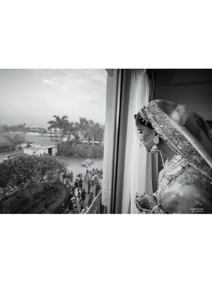 A powerful black and white shot of the bride looking out the window at her own baraat arriving, a moment filled with anticipation.