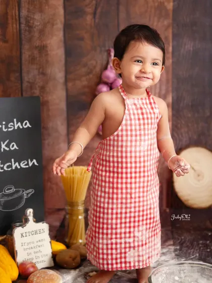 This little chef is all smiles in her gingham apron. The 'Prisha Ka Kitchen' setup is ready for some fun and messy cake smashing.