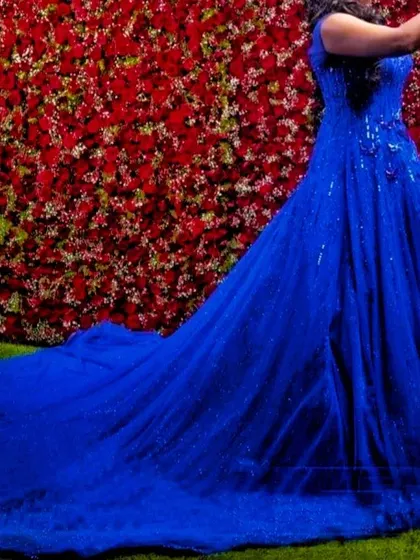 The long, sparkling trail of our royal blue ball gown is on full display against a vibrant red flower wall.