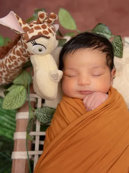 A close-up of a peacefully sleeping newborn in a warm, mustard-colored wrap, snuggled next to a friendly giraffe toy. The earthy tones and jungle-like foliage add a touch of adventure.
