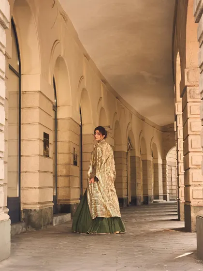 This wide shot shows the full effect of the modern lehenga with its flowing green skirt and longline striped jacket, set against a beautiful architectural backdrop.