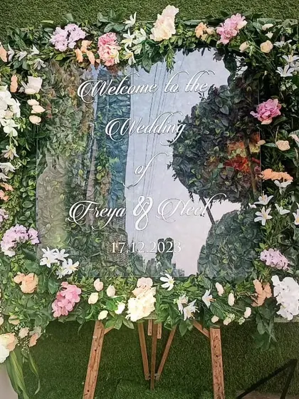 An elegant and modern welcome sign for a wedding. The message is written on a clear acrylic board, which is framed with beautiful pastel-colored flowers for a fresh and sophisticated look.