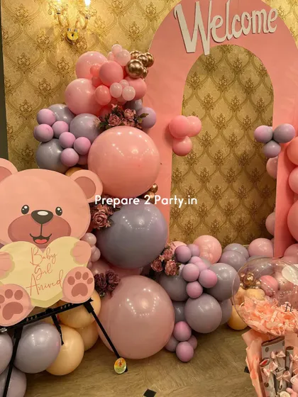A detailed shot of the welcome decoration, focusing on a cute pink teddy bear cutout holding a "Baby Girl Arrived" sign. The surrounding balloons in pastel pink and purple add to the charm.