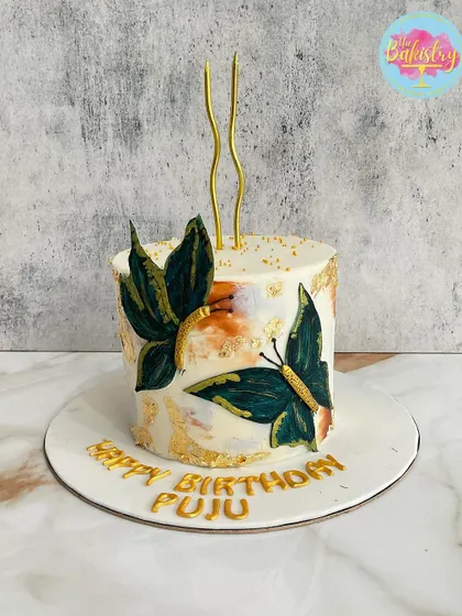 A modern, artistic cake with hand-painted green butterflies and gold leaf accents for "Puju's" birthday.