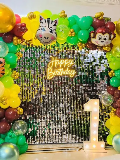 A first birthday party setup with a jungle theme against a shimmer wall. This decoration combines a sparkling backdrop with a colorful balloon arch, animal foil balloons, and a light-up number '1'.