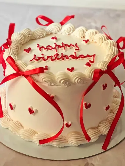 A classic white and red ribbon cake, perfect for birthdays or anniversaries. The simple design with piped borders and tiny hearts is effortlessly charming.