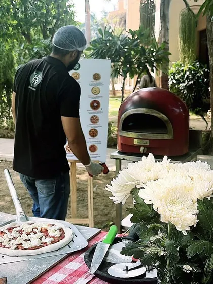 Setting up for an outdoor catering event. My chef stands ready with a pizza on the peel, the red wood-fired oven in the background, and a menu board displaying the day's options for guests.