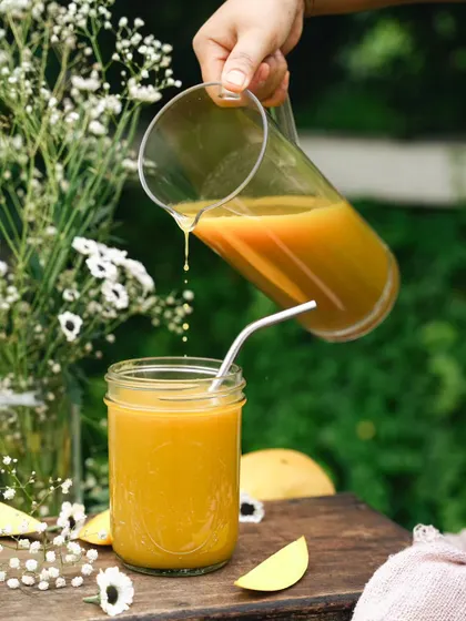 A beautiful outdoor shot of fresh mango juice being poured. The natural garden setting and soft light give this image a wholesome, picnic-like feel.